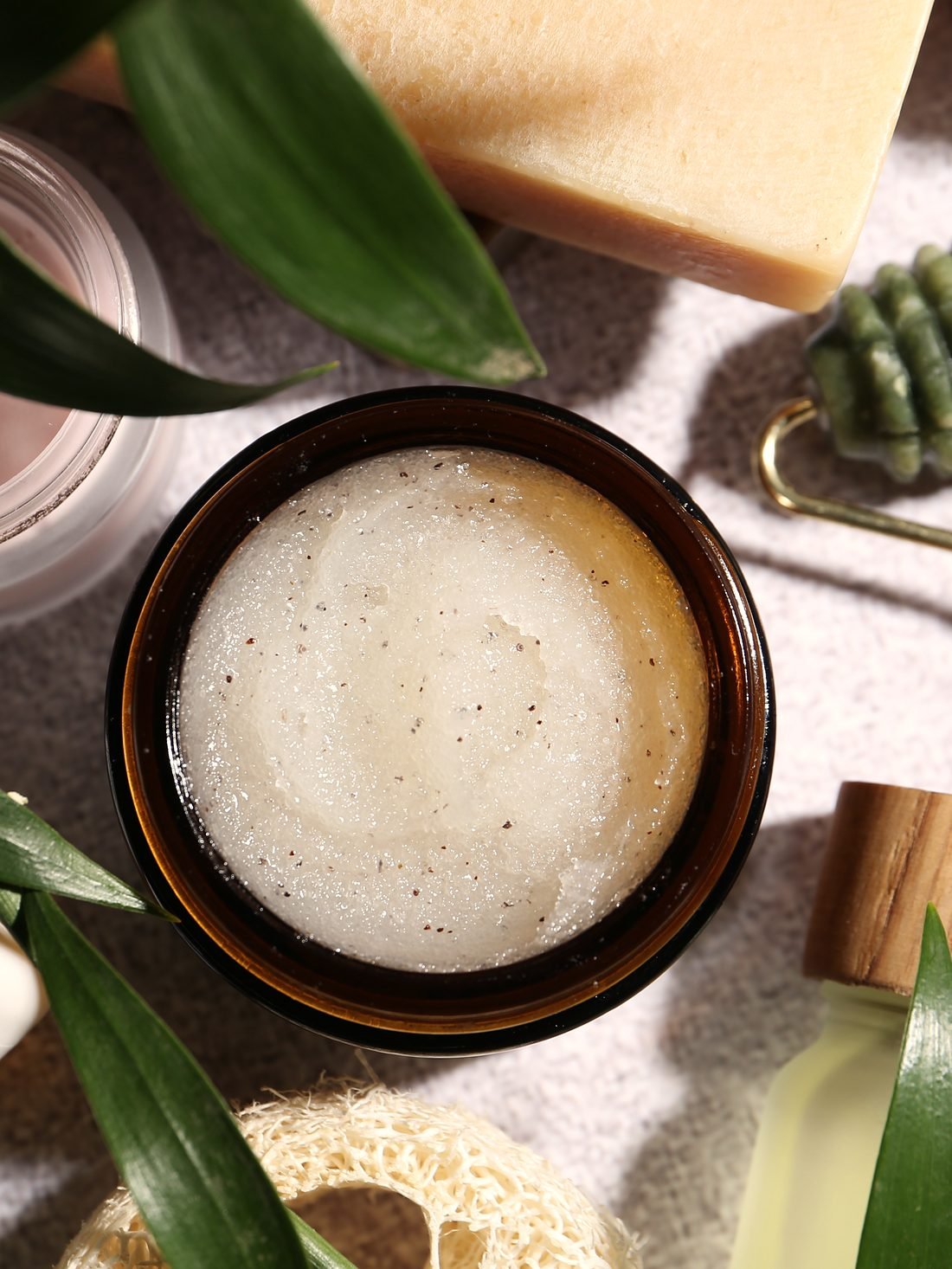 A jar of scrub, a jade facial roller and skincare bottles among green leaves